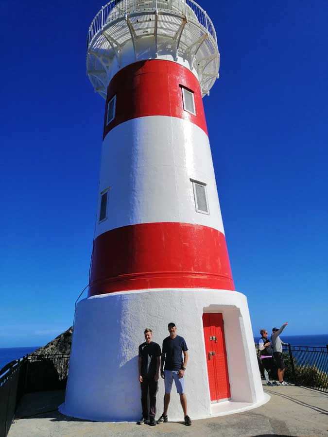 two men stand in front of Cape Palliser lighthouse on a sunny day