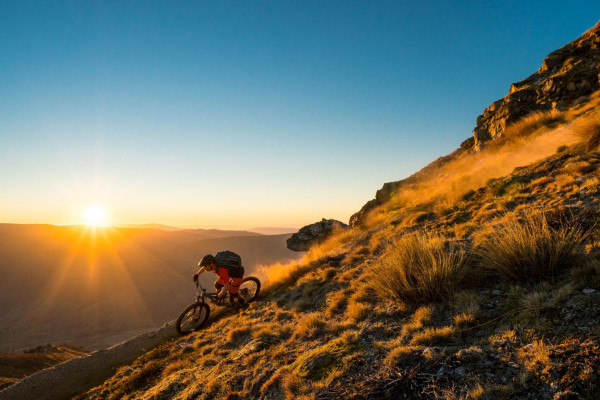 A mountain biker rides down a dusty trail with the glowing sun as a backdrop.